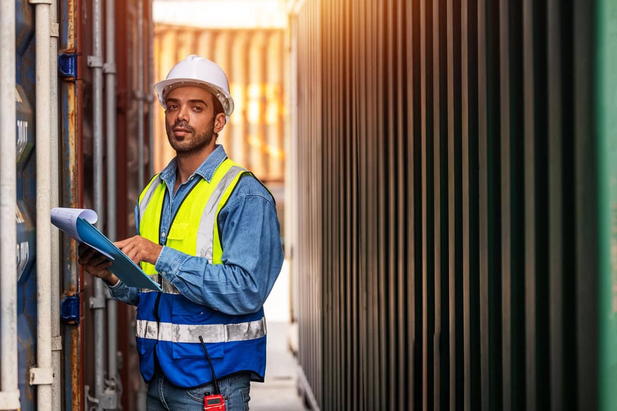 internal-communications-for-transport-and-logistics-commskit A male frontline transport and logistics worker wearing PPE and standing in front of a shipping container representing internal communications for transport and logistics by CommsKit